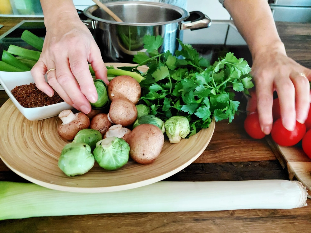 NaturTeller – vegane Ernährungsberatung - Zwei Hände bereiten frisches Gemüse auf einer Holztheke vor. Eine Hand greift in eine flache Holzschale mit Rosenkohl und Champignons, während die andere Hand sich einem Haufen leuchtend roter Tomaten nähert.