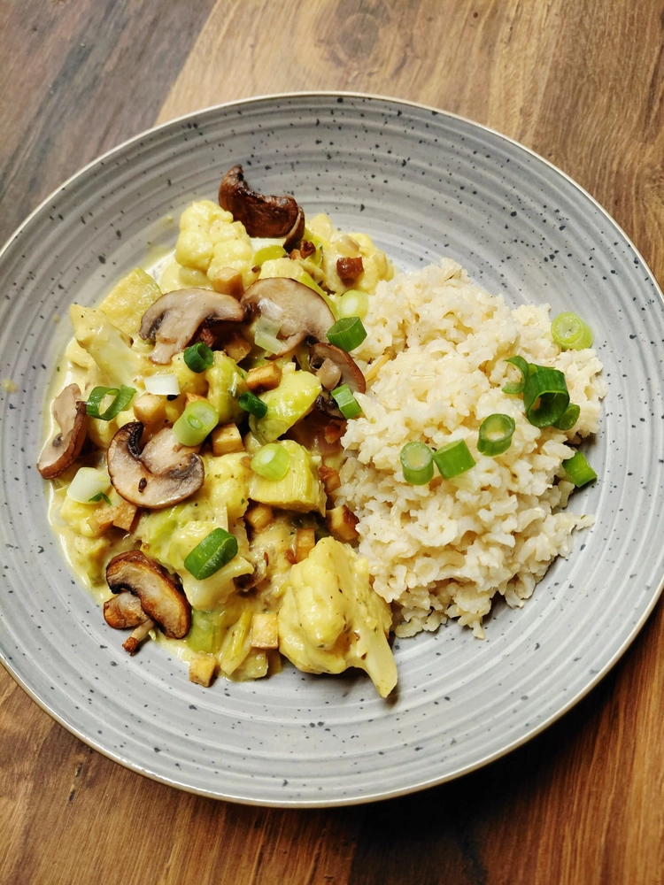 NaturTeller – vegane Ernährungsberatung - Eine helle, flache Schüssel mit einem cremigen Gemüsecurry, serviert mit Reis. Das Curry links enthält Blumenkohlröschen, in Scheiben geschnittene braune Champignons und Frühlingszwiebeln in einer gelblichen Soße. Rechts befindet sich ein Berg weißen Reis, ebenfalls mit gehackten Frühlingszwiebeln garniert.