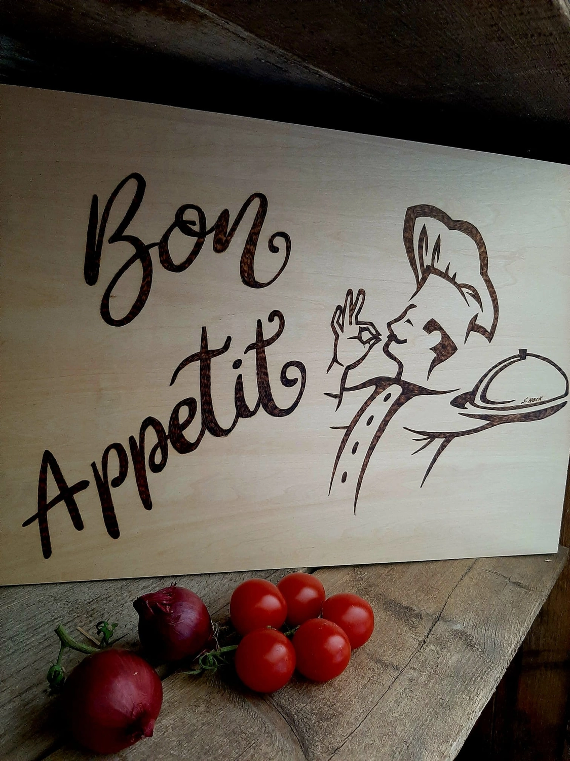 NaturTeller – vegane Ernährungsberatung - Ein eingebranntes Holzschild mit der Aufschrift „Bon Appetit“ in Schreibschrift. Es zeigt die Illustration eines Kochs mit traditioneller Kochmütze, der eine abgedeckte Servierplatte hält. Vor dem Schild liegen einige frische Kirschtomaten auf der Theke.