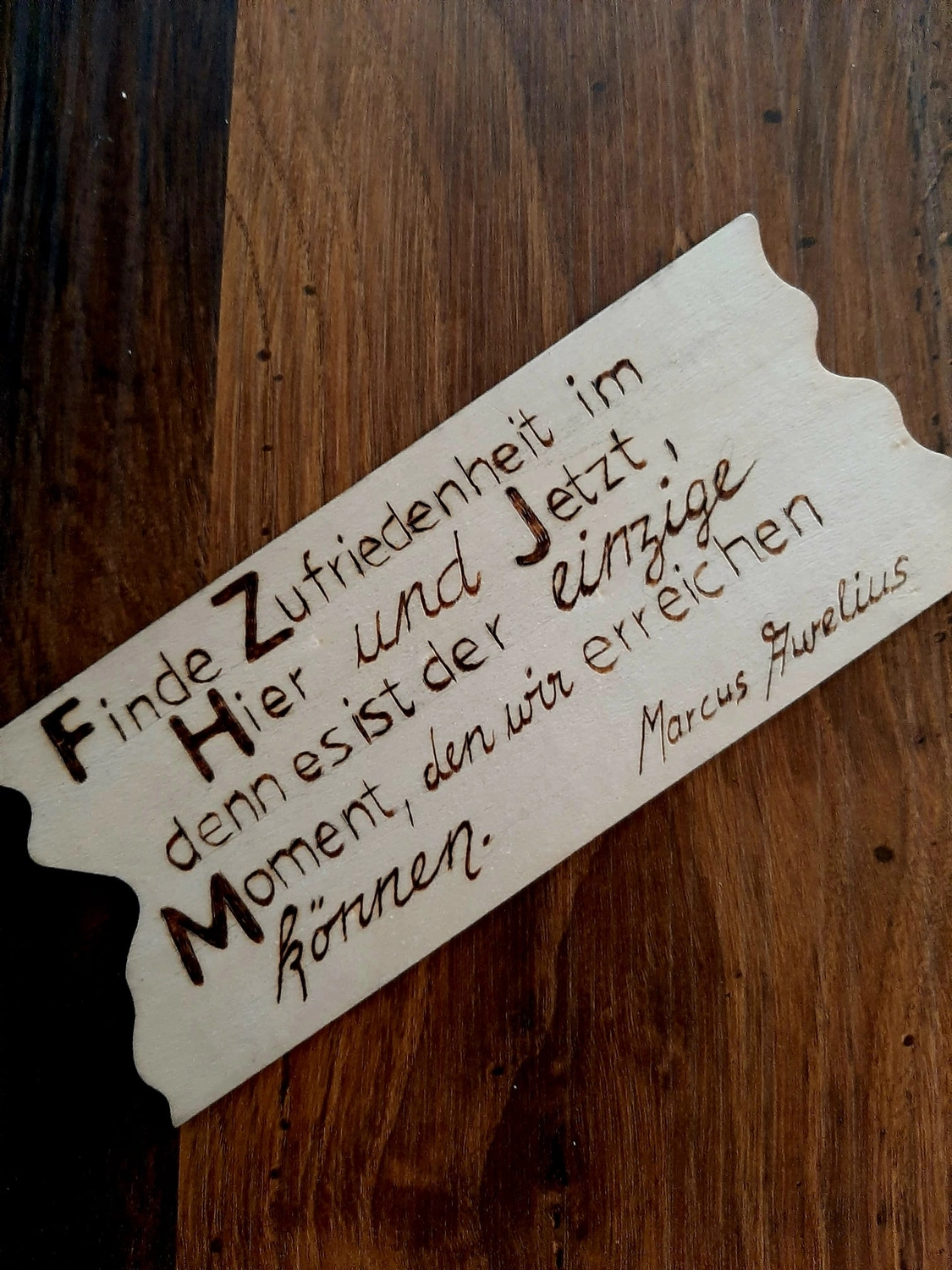 NaturTeller – vegane Ernährungsberatung - Ein Stück helles Holz mit rauen, „ausgerissenen“ Kanten, das an Pergament erinnert. Darauf ist ein deutsches Zitat eingebranntes Zitat, das besagt, dass man im „Hier und Jetzt“ Zufriedenheit finden kann, da dies der einzige Moment ist, den wir wirklich erreichen können.