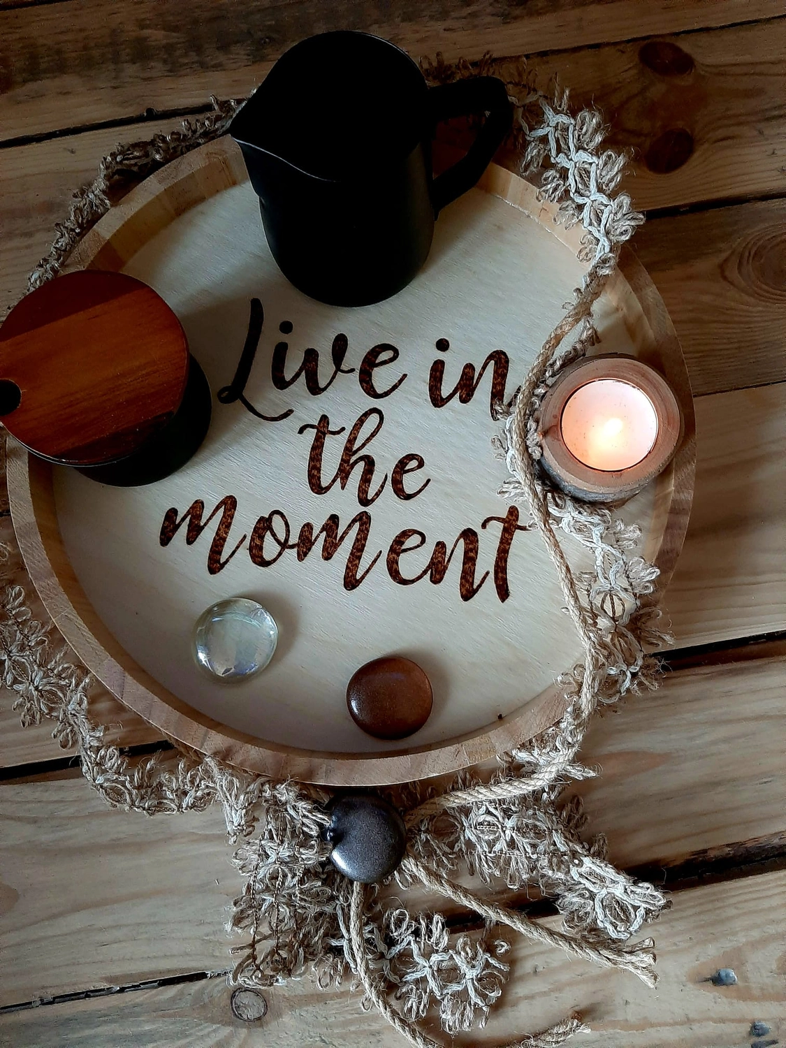 NaturTeller – vegane Ernährungsberatung - Eine rustikale, runde Holzscheibe, die als dekoratives Tablett dient. Sie trägt die eingebrannte Phrase „Lebe im Moment“ und steht auf einem Stück Jute. Auf dem Tablett befinden sich ein kleiner dunkler Krug, ein Teelicht in einem Glashalter und mehrere glatte Ziersteine.
