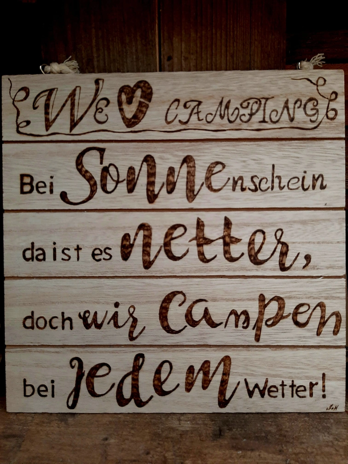 NaturTeller – vegane Ernährungsberatung - Ein Holzschild aus horizontalen Latten mit einem eingebrannten deutschen Gedicht über das Campen. Es beginnt mit „Wir ❤️ Camping“ und bedeutet so viel wie: „Bei Sonnenschein ist es schöner, aber wir zelten bei jedem Wetter!“